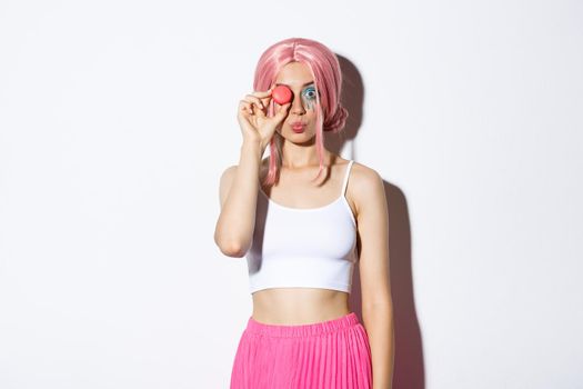 Portrait of lovely girl in pink wig, dressed up for party or holiday celebration, holding delicious macaroon and pouting silly, standing over white background.