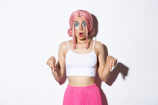 Shocked caucasian girl with pink wig and bright makeup gasping, looking scared and pointing fingers down at halloween banner, standing over white background.