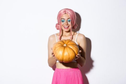 Portrait of cheerful woman with pink wig and bright makeup, looking happy at pumpkin for halloween, standing over white background.