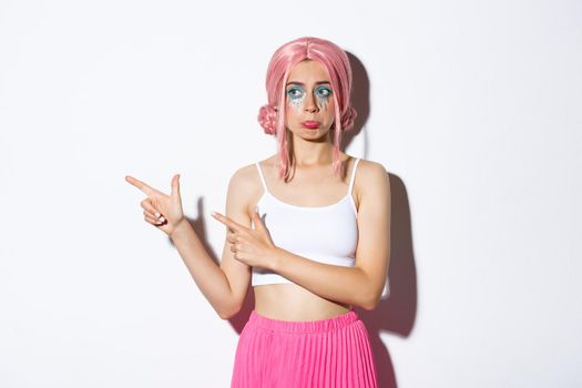 Image of sad cute girl with pink wig and halloween makeup, sulking upset and frowning, pointing fingers left at something disappointing, standing over white background.