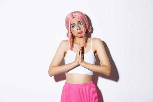 Portrait of silly sad girl in pink anime wig asking for help, holding hands clasped together in pray, begging for something with cute pouting expression, standing over white background.