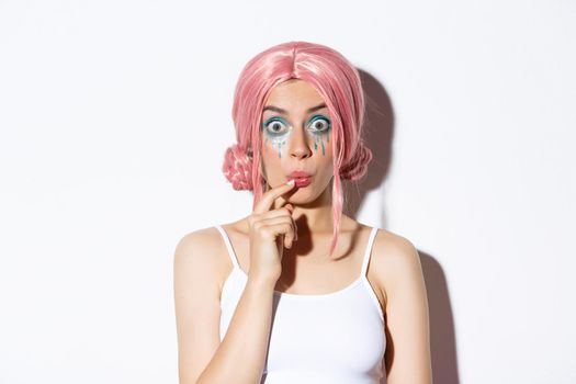 Close-up of silly pretty girl in pink wig, looking thoughtful at camera, standing over white background.