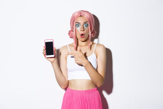 Portrait of surprised beautiful girl in halloween costume, pink wig and bright makeup, pointing finger at smartphone screen, standing over white background.