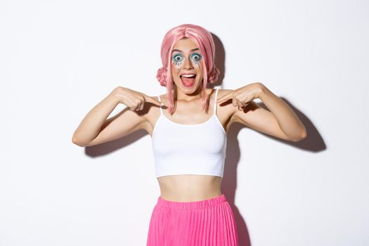 Portrait of surprised party girl with pink wig and bright makeup, pointing at herself and smiling excited, showing her halloween outfit, standing over white background.