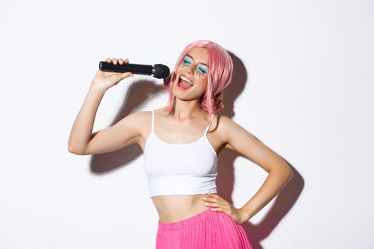 Portrait of joyful beautiful female in pink wig, with bright makeup, singing karaoke in microphone, standing over white background.