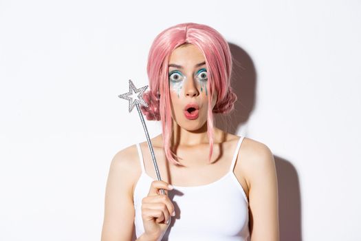 Close-up of surprised female model in pink wig and fairy makeup, holding magic wand and looking amazed at camera, standing over white background.