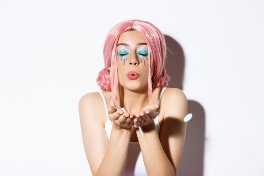 Close-up of lovely pretty party girl with pink wig and bright makeup, blowing confetti from palm, celebrating holiday, standing over white background.