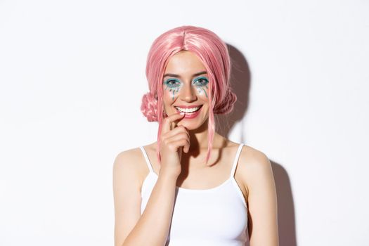 Close-up of coquettish smiling woman in pink party wig, touching lip and looking with temptation, standing over white background.