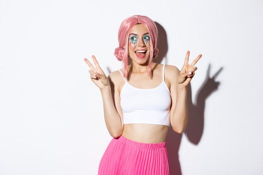 Portrait of stylish attractive girl in pink wig and halloween costume, showing peace signs and smiling happy, standing over white background.