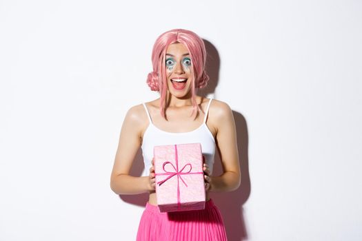 Portrait of surprised attractive girl looking excited, receive gift for birthday, wearing pink wig, standing over white background.
