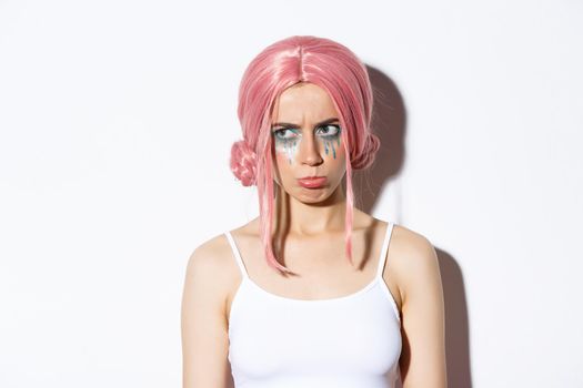 Close-up of cute girl with pink hair and glamour makeup, sulking and looking jealous left, feeling offended, standing over white background.