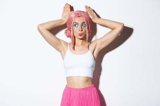 Portrait of lovely party girl in pink anime wig, showing bunny ears and pouting silly, celebrating halloween, standing over white background.