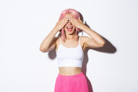 Portrait of amused cute party girl with pink hair close eyes with hands and waiting for surprise, smiling happy, standing over white background.