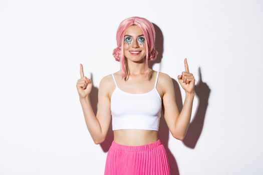 Portrait of happy smiling female model with pink anime wig and halloween makeup, looking satisfied at banner, pointing fingers up, standing over white background.