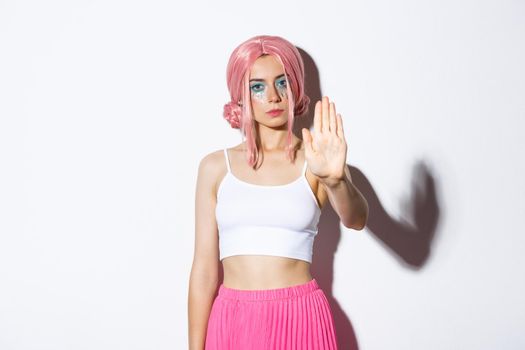 Image of serious female model in pink wig and halloween costume showing stop sign, extend hand forward to reject or prohibit something bad, standing over white background.