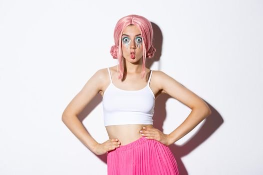 Image of silly party girl with pink wig and bright makeup, celebrating halloween, looking surprised, standing over white background.