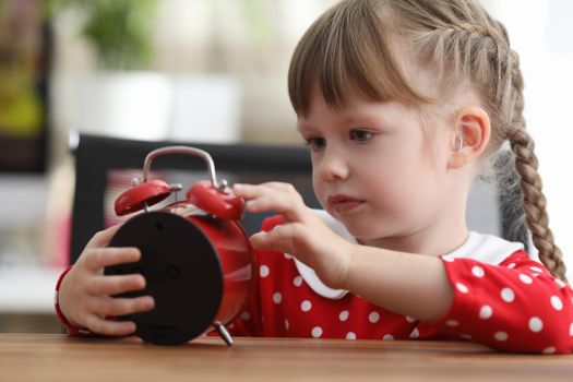 Little girl with pigtails looking at red alarm clock at table. Lesson schedule for kids concept