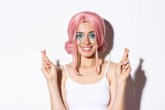 Close-up of lovely smiling woman in halloween costume, pink wig and bright makeup, looking hopeful at camera and making wish with fingers crossed.
