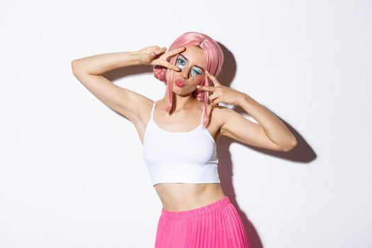 Carefree stylish girl with pink wig and bright makeup, having fun on party, celebrating and dancing, showing peace signs, standing over white background.