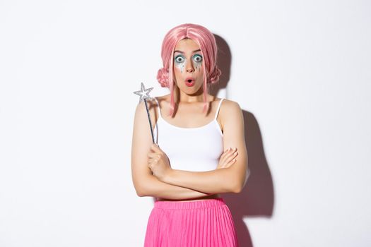 Portrait of surprised girl in pink wig, dressed as fairy for halloween party, holding magic wand and looking wondered, standing over white background.