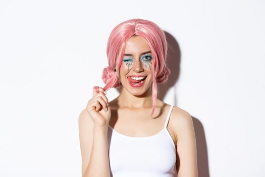 Portrait of sassy attractive girl in pink wig, wearing outfit for halloween party, smiling and winking at camera, standing over white background.