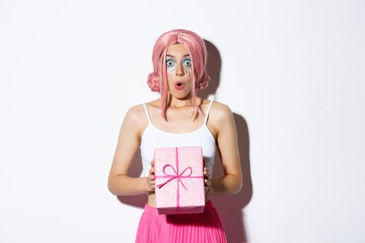 Portrait of surprised attractive girl looking excited, receive gift for birthday, wearing pink wig, standing over white background.