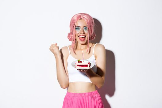 Portrait of cheerful smiling girl celebrating her birthday, wearing pink wig, holding b-day cake and shouting of joy, standing over white background.