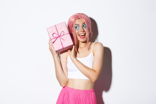 Image of beautiful girl in pink wig shaking box with birthday gift, wonder what inside wrapped box, standing over white background.