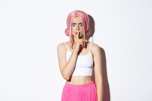 Image of angry frowning girl in pink wig and halloween makeup, shushing at someone, scolding person for being too loud, standing over white background.