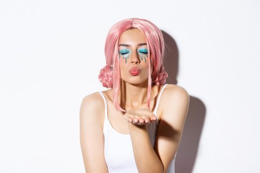 Close-up of lovely girl in halloween costume with pink wig and bright makeup, close eyes and blowing air kiss at camera, standing over white background.