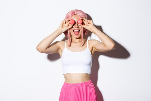 Portrait of beautiful party girl in pink wig, holding macaroons over eyes and smiling amazed, standing over white background.