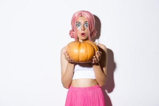 Image of attractive girl in pink wig looking amazed while holding pumpkin for halloween party, standing over white background.