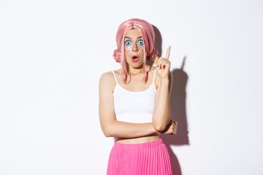 Image of creative woman in pink party wig and bright makeup, suggesting idea, raising index finger in eureka sign, standing over white background.