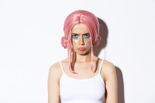 Close-up of angry cute girl with pink wig and glamour makeup, sulking mad at someone, feeling offended, standing over white background.