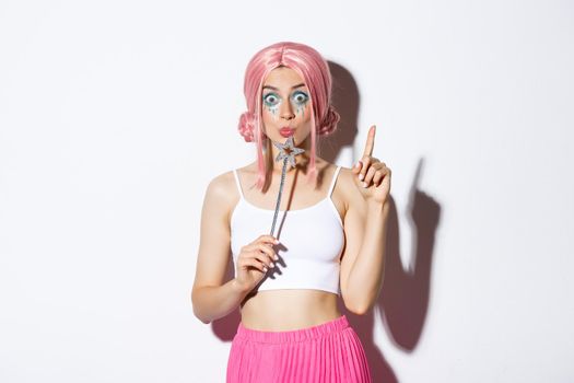Portrait of beautiful girl in fairy costume looking thoughtful, holding magic wand and wearing pink wig, have an idea, standing over white background.