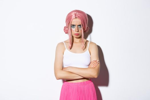 Portrait of angry cute girl with pink wig and glamour makeup, cross arms on chest and sulking mad at someone, feeling offended, standing over white background.