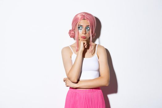 Portrait of thoughtful sad girl with pink hair and bright makeup, wearing halloween outfit, looking left and pouting upset, standing over white background.