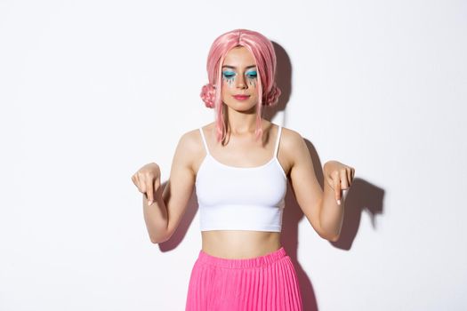 Portrait of smiling party girl in pink wig and outfit for halloween, pointing and looking down at your logo, standing over white background.