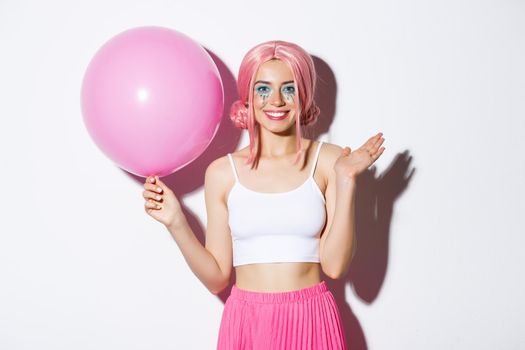 Image of cheerful young girl with pink wig and bright makeup, holding balloon and waving hand to say hi, greeting someone at party, celebrating holiday, white background.