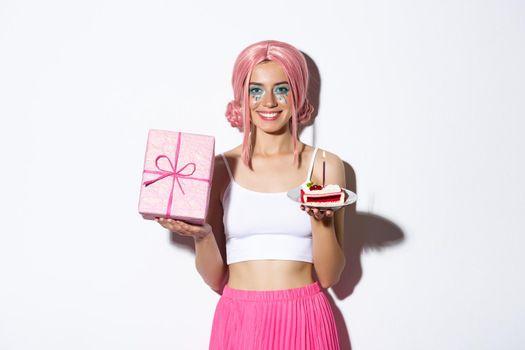 Beautiful girl celebrating birthday in pink wig, holding gift and b-day cake, standing over white background.