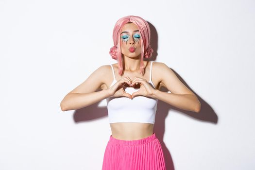 Silly beautiful female model in pink wig, with glamour makeup, showing heart gesture and kissing, express sympathy or love, standing over white background.
