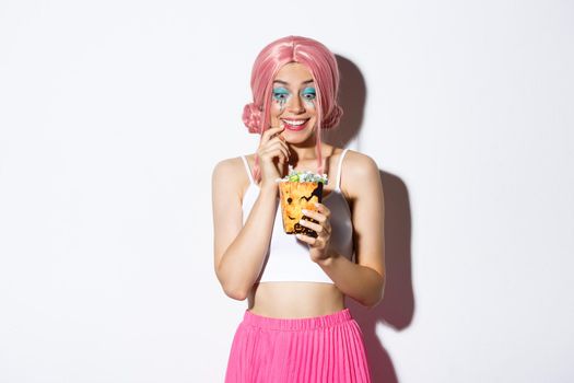 Portrait of excited beautiful girl celebrating halloween, looking at sweets with tempted expression, trick or treating in pink wig, standing over white background.