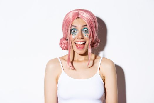 Close-up of surprised happy girl in pink wig, smiling and looking excited at camera, standing over white background, celebrating halloween.