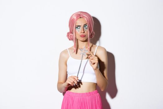 Image of indecisive pouting girl in fairy costume, holding magic wand, looking thoughtful and standing over white background.