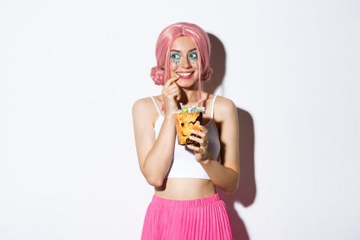 Portrait of thoughtful smiling girl in pink wig, thinking about trick or treat while holding candies, celebrating halloween, standing over white background.