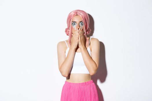 Portrait of shocked woman in pink party wig looking ambushed, gasping and covering mouth with hands, seeing something shocking or scary, wearing halloween costume.
