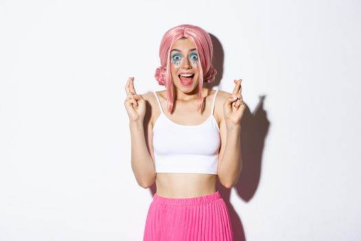 Image of excited party girl crossing fingers for good luck and smiling amazed, looking hopeful, wearing halloween costume with pink wig and bright makeup.