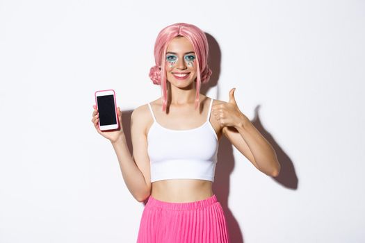 Image of attractive young party girl in pink wig, with bright makeup, showing thumbs-up and mobile phone screen, recommend app, standing over white background.