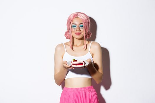 Image of beautiful girl in pink wig close eyes and smelling tasty cake, celebrating holiday, standing over white background.