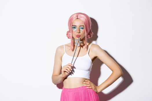 Image of silly attractive girl in pink wig, dressing as fairy for halloween party, hoding magic wand, standing over white background.